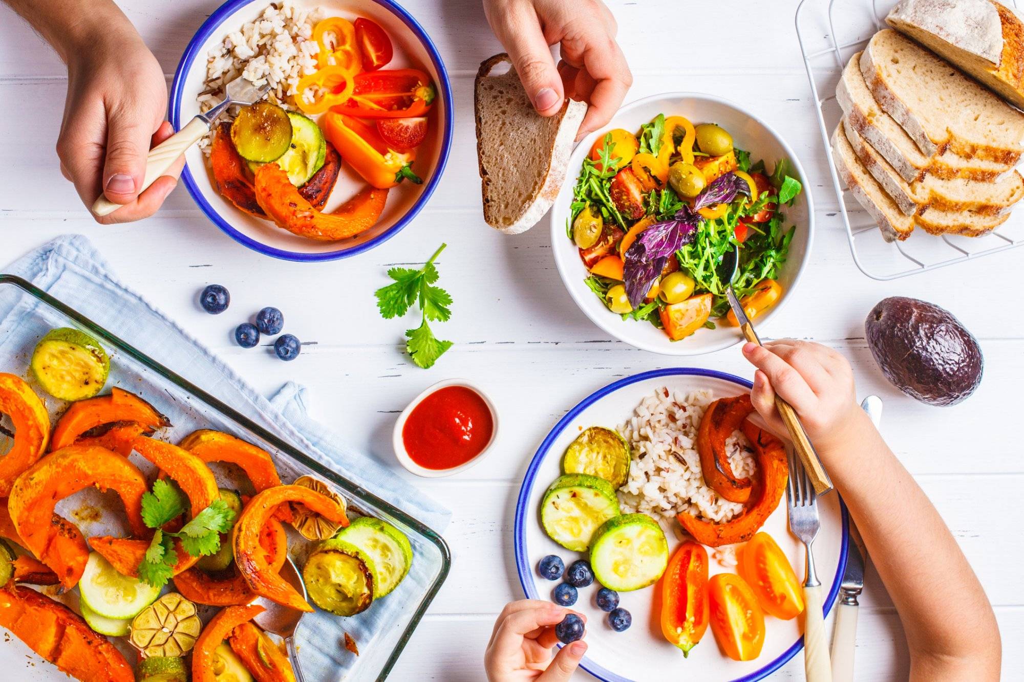 Family eating a healthy vegetarian food. Vegan lunch table top view, plant based diet. Baked vegetables, fresh salad, berries, bread on a white background.