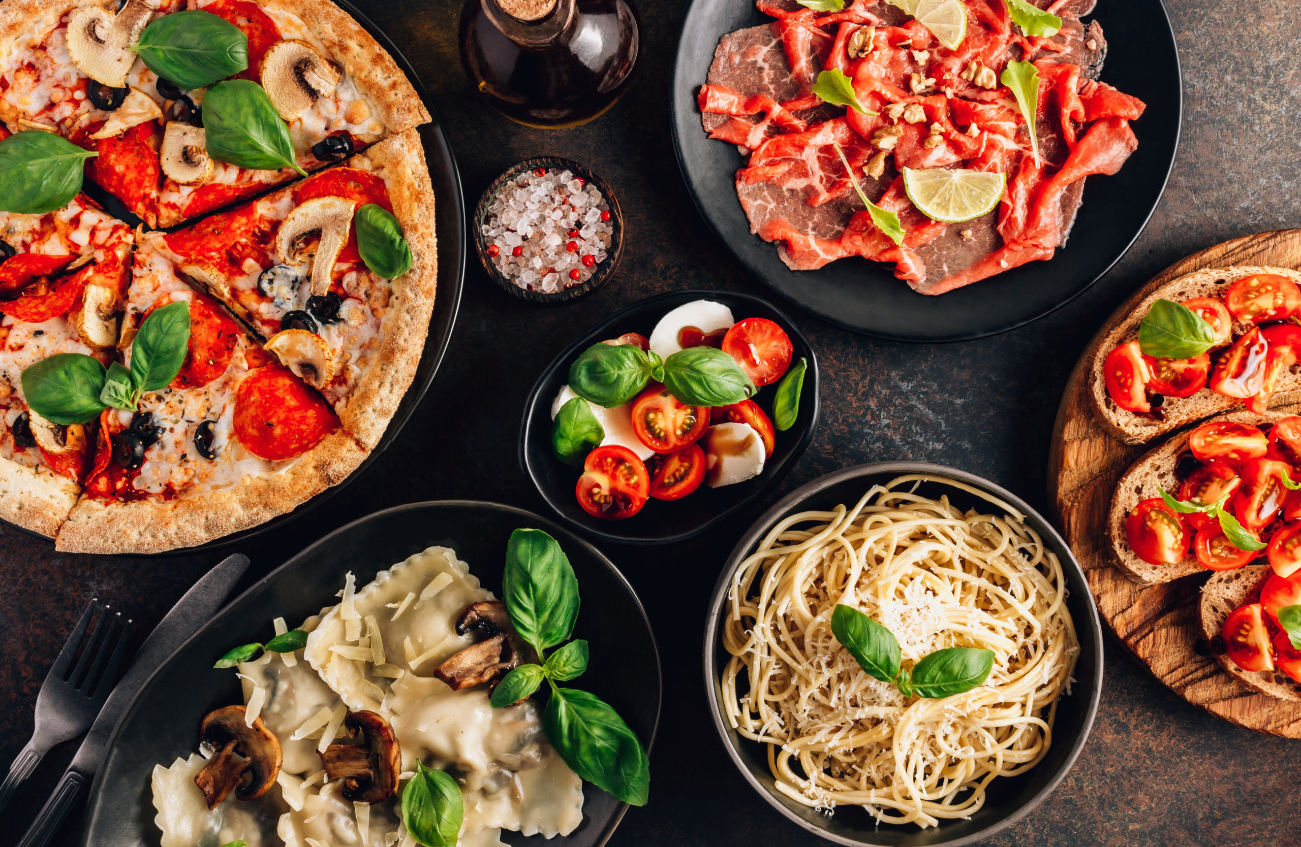 Full table of italian meals on plates Pizza, pasta, ravioli, carpaccio. caprese salad and tomato bruschetta on black background.