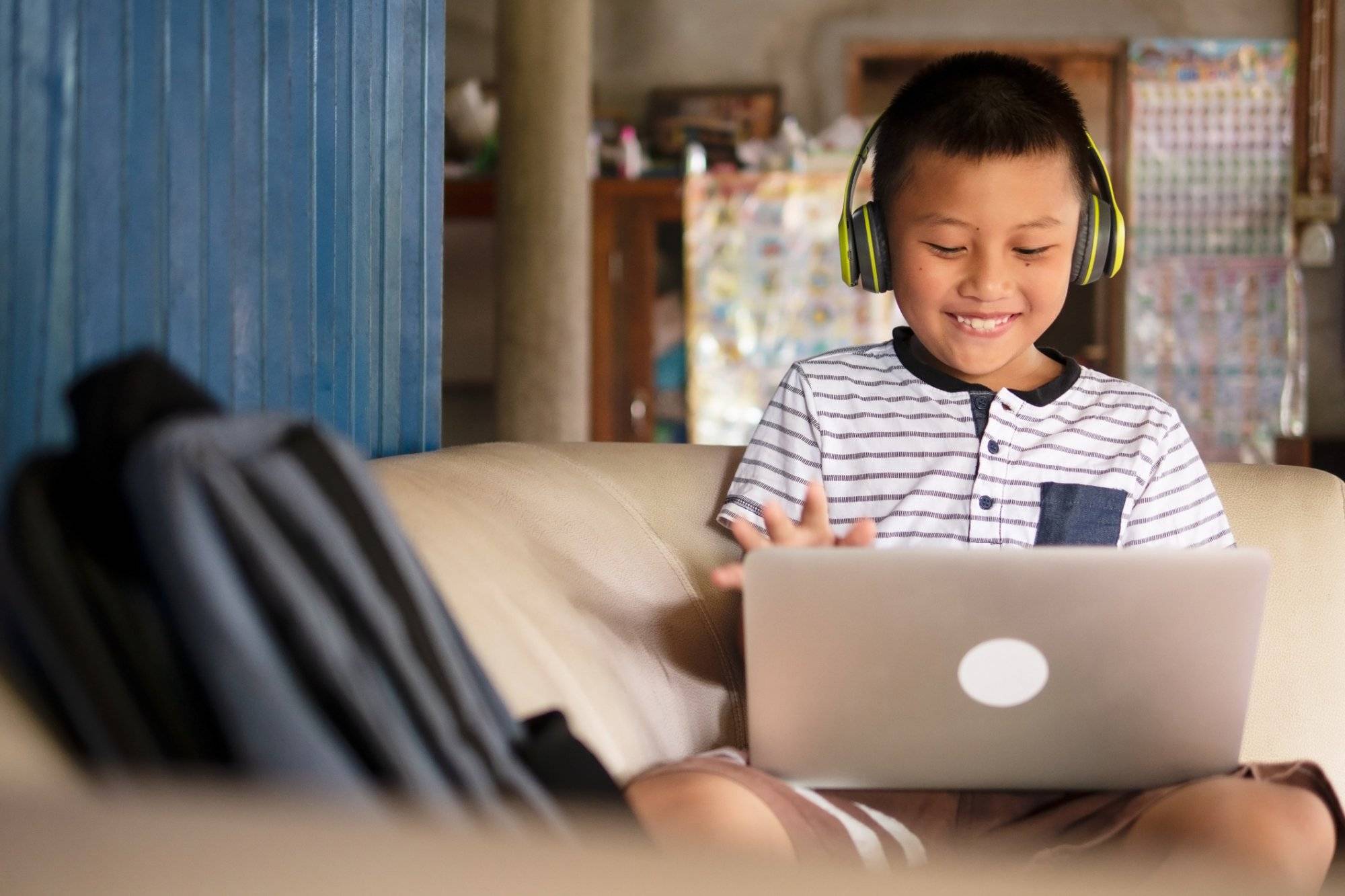 Online remote learning, distance education and homeschooling concepts. School kid Asian preteen boy in headphone using laptop computer on couch in rustic rural home during COVID-19 pandemic.