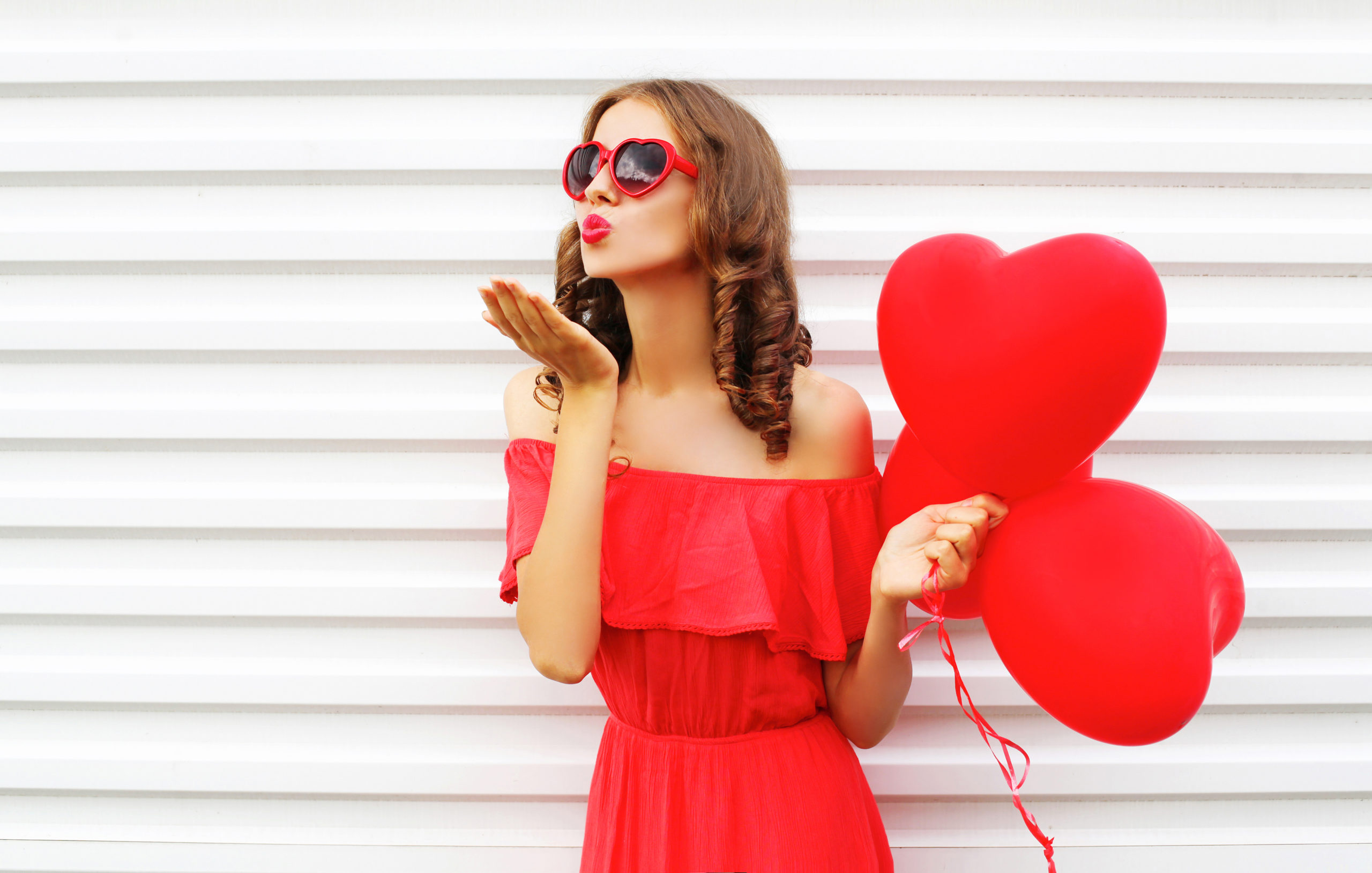 Portrait woman in red dress sends air kiss with balloon heart shape over white background